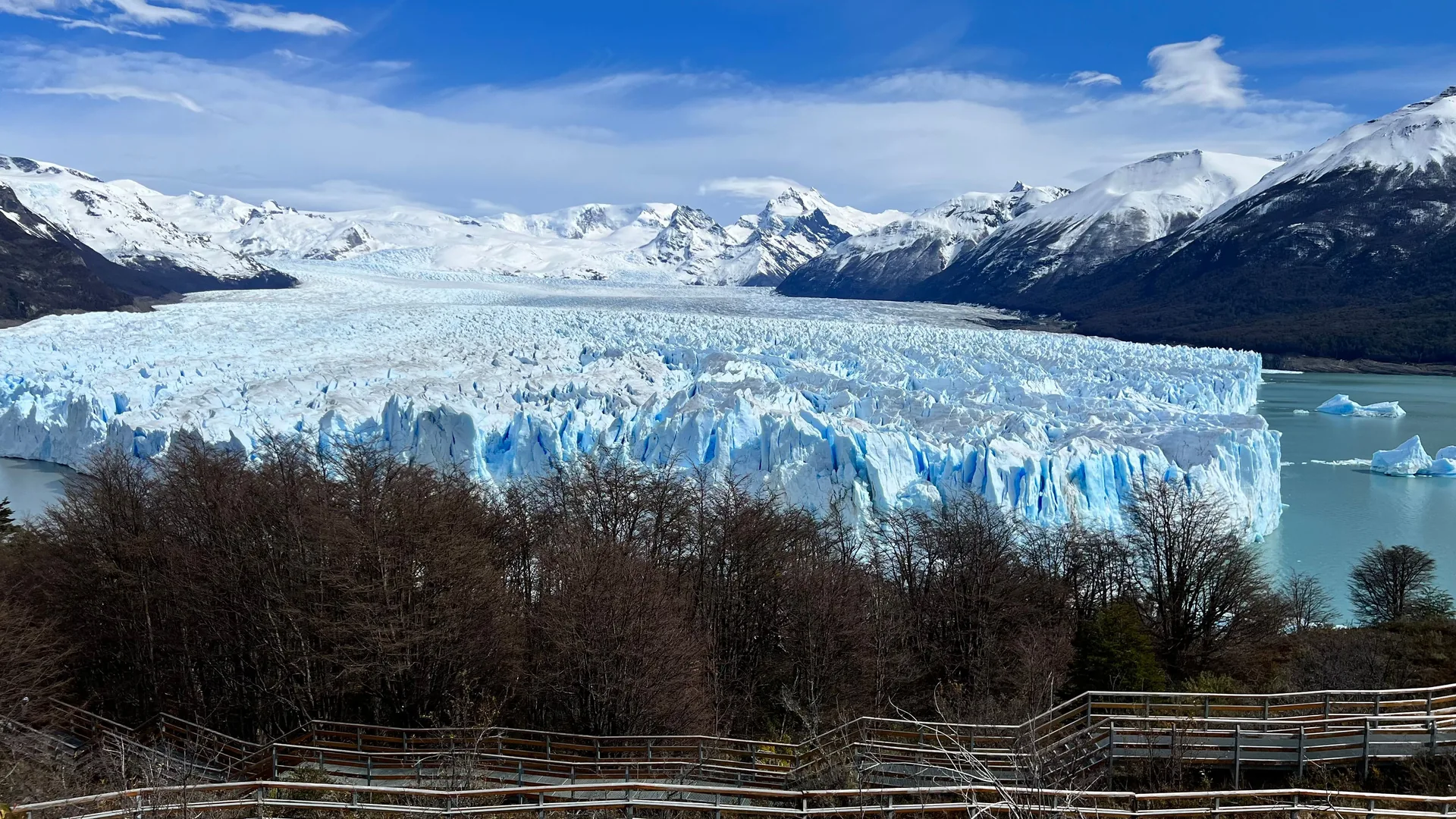 Široký pohled na zubaté modrobílé čelo Ledovce Perito Moreno nad jezerem, se zasněženými vrcholy v pozadí